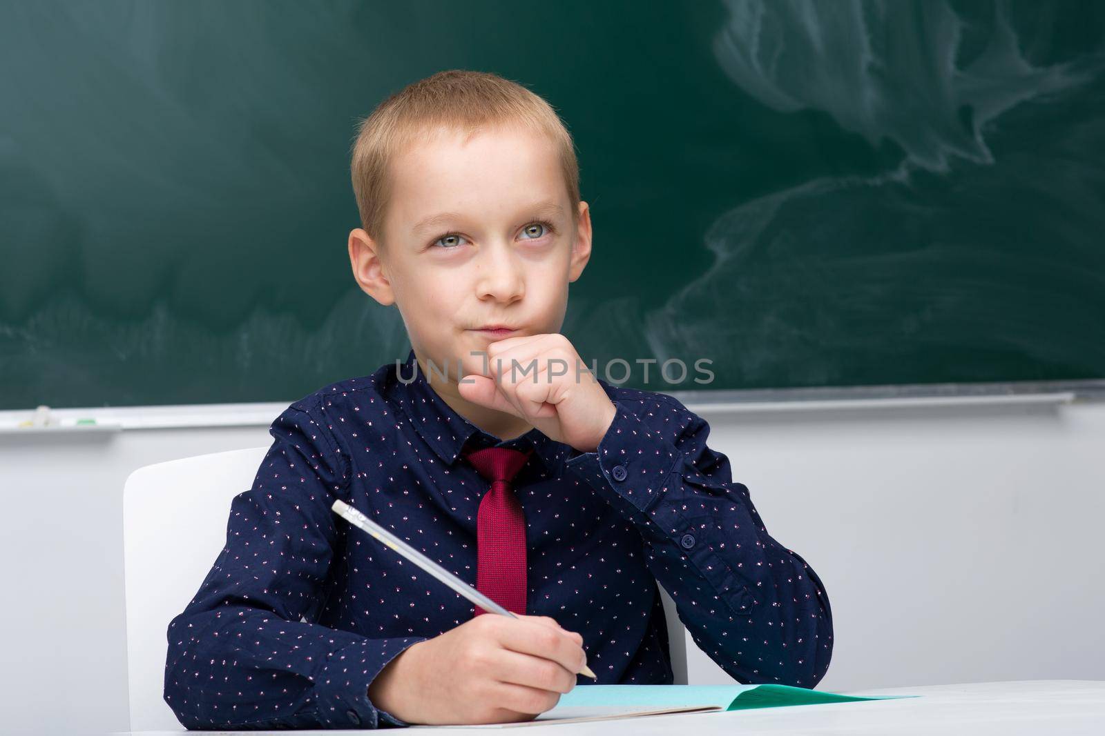 Thinking schoolboy studying at school. Boy student in blue shirt and necktie sitting at desk on background of blackboard in classroom. Back to school, education concept