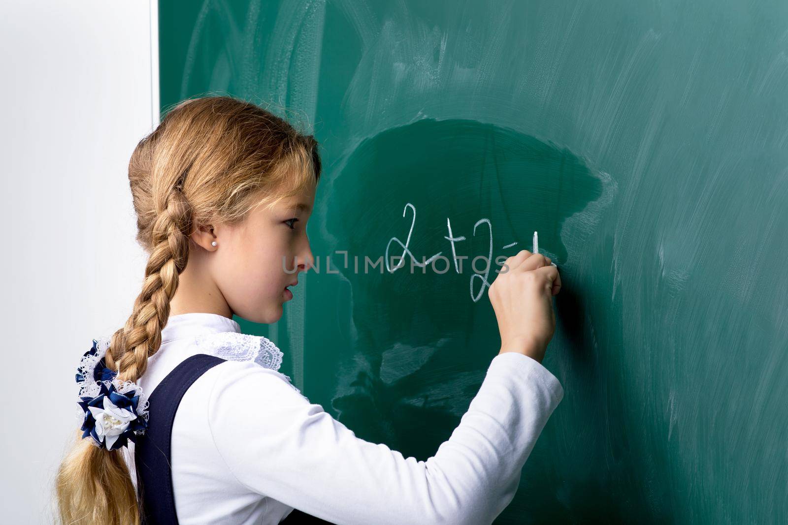 Schoolgirl writing on chalkboard in classroom. Primary school student in uniform standing in front of blackboard and writing using chalk. Back to school, education concept
