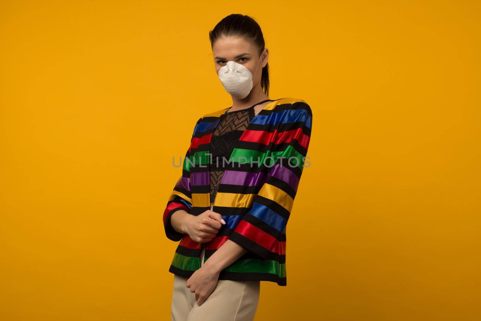 Beautiful slim girl fashion model posing in a protective respirator on a yellow background. LGBT community rainbow color jacket