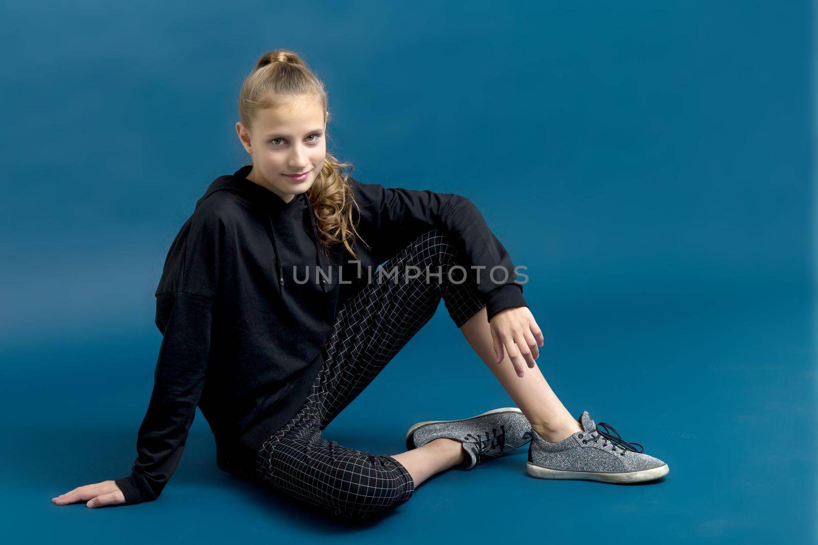 Teen girl in black hoodie sitting on floor. Portrait of beautiful serious teenager dressed black sweatshirt, breeches and sneakers posing against blue background in studio