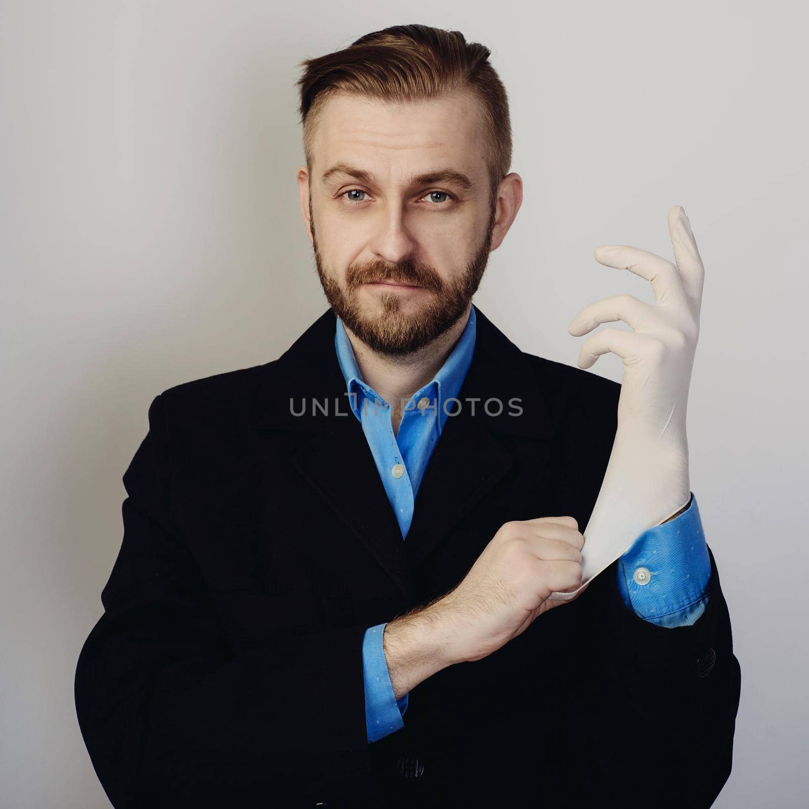 attractive stylish bearded businessman in suit and blue shirt puts on medical glove on gray background