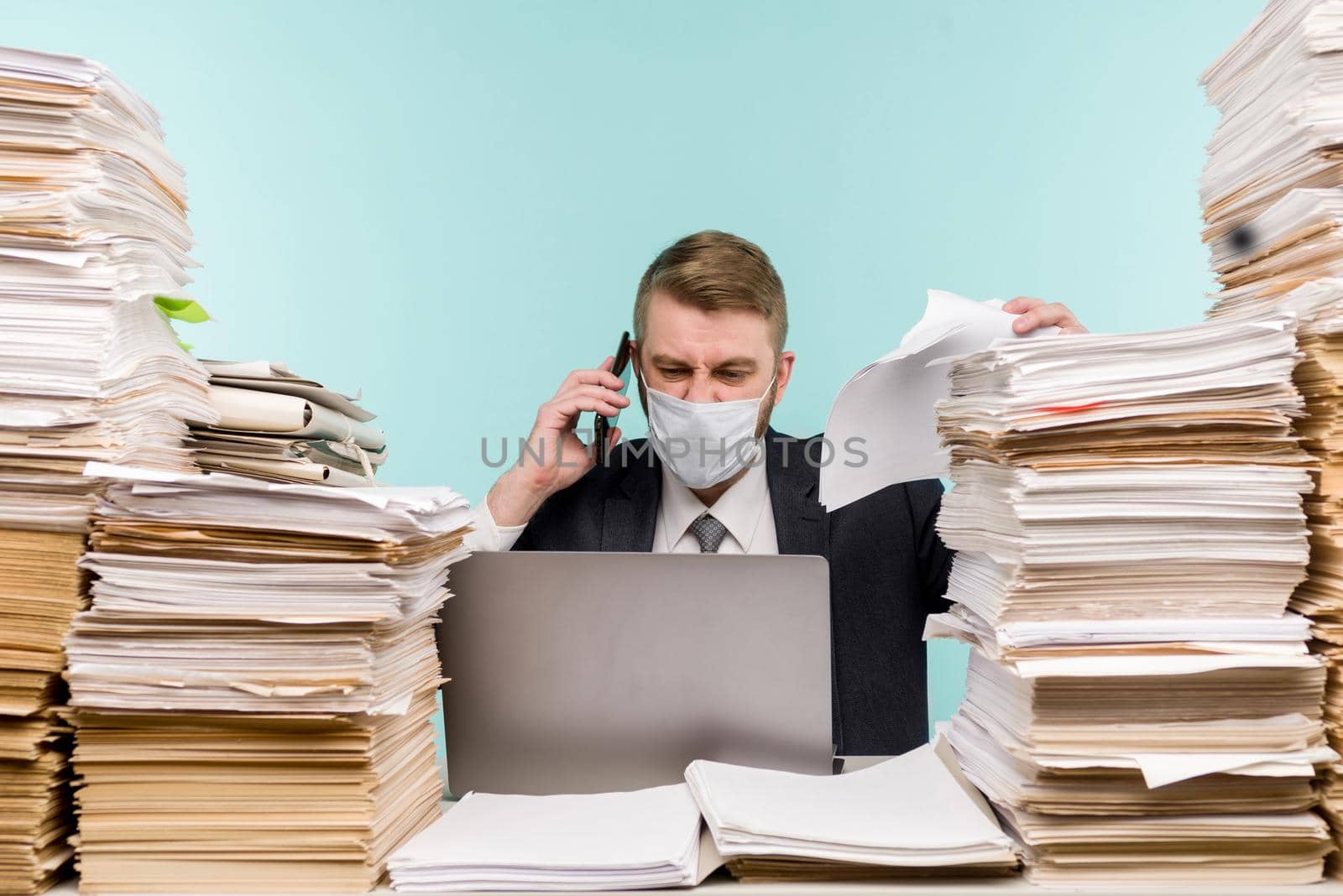 A male accountant or company manager works in an office in a pandemic in view of the accumulated paper work. A protective medical mask is on the face. On the desktop are large stacks of documents. - image