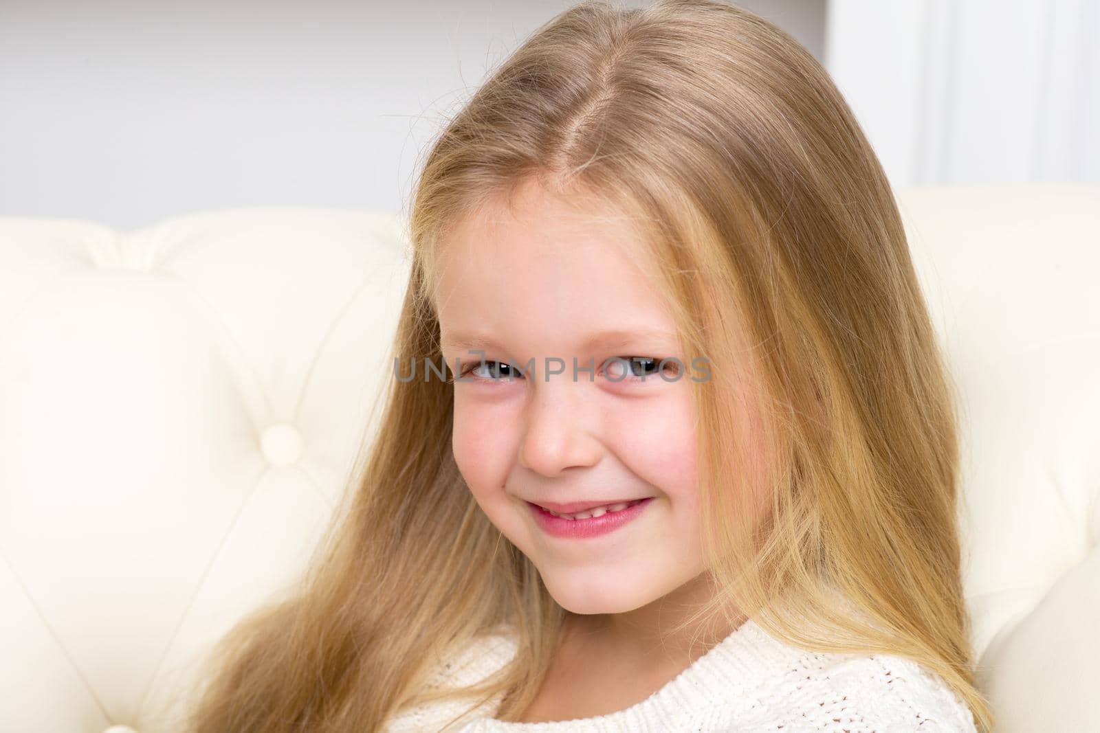 Close up portrait of lovely blonde girl sitting on comfortable sofa. Cute smiling long haired little girl having fun at home. Adorable happy child resting on couch