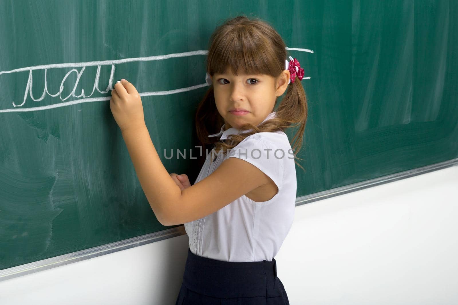 Serious schoolgirl standing at blackboard. Portrait of elementary school student girl in white blouse writing on green chalkboard in classroom and looking at camera. School and education concept