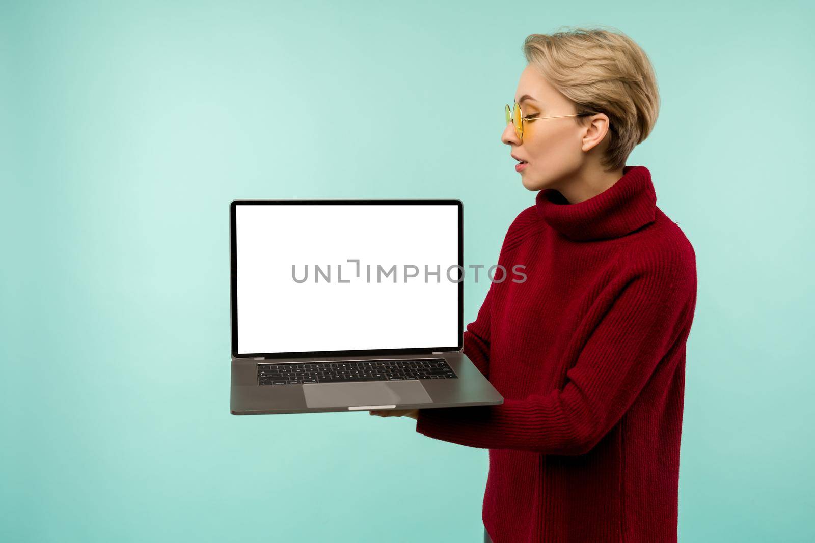 Young girl in a red sweater demonstrates a blank laptop screen on a blue background - image