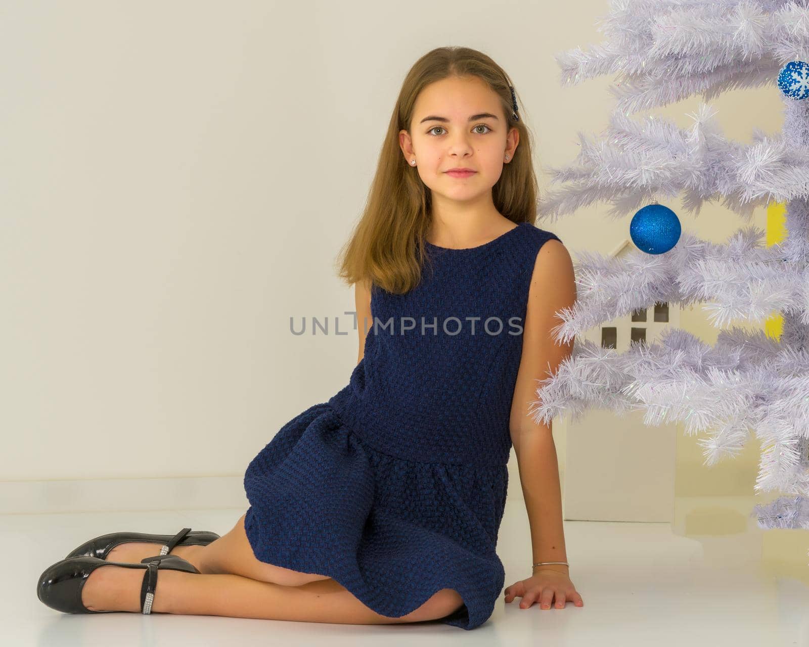 Beautiful Blonde Girl Wearing Stylish Dress Sitting on the Floor in Front of Decorated White Christmas Tree, Adorable Girl Celebrating Christmas and New Year Smiling at Camera