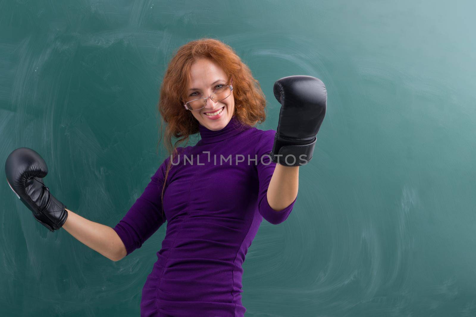 Smiling teacher wearing black boxing gloves celebrating her winning in front of blank green chalkboard. Young woman wearing glasses and purple casual dress standing in winning pose with arms raised