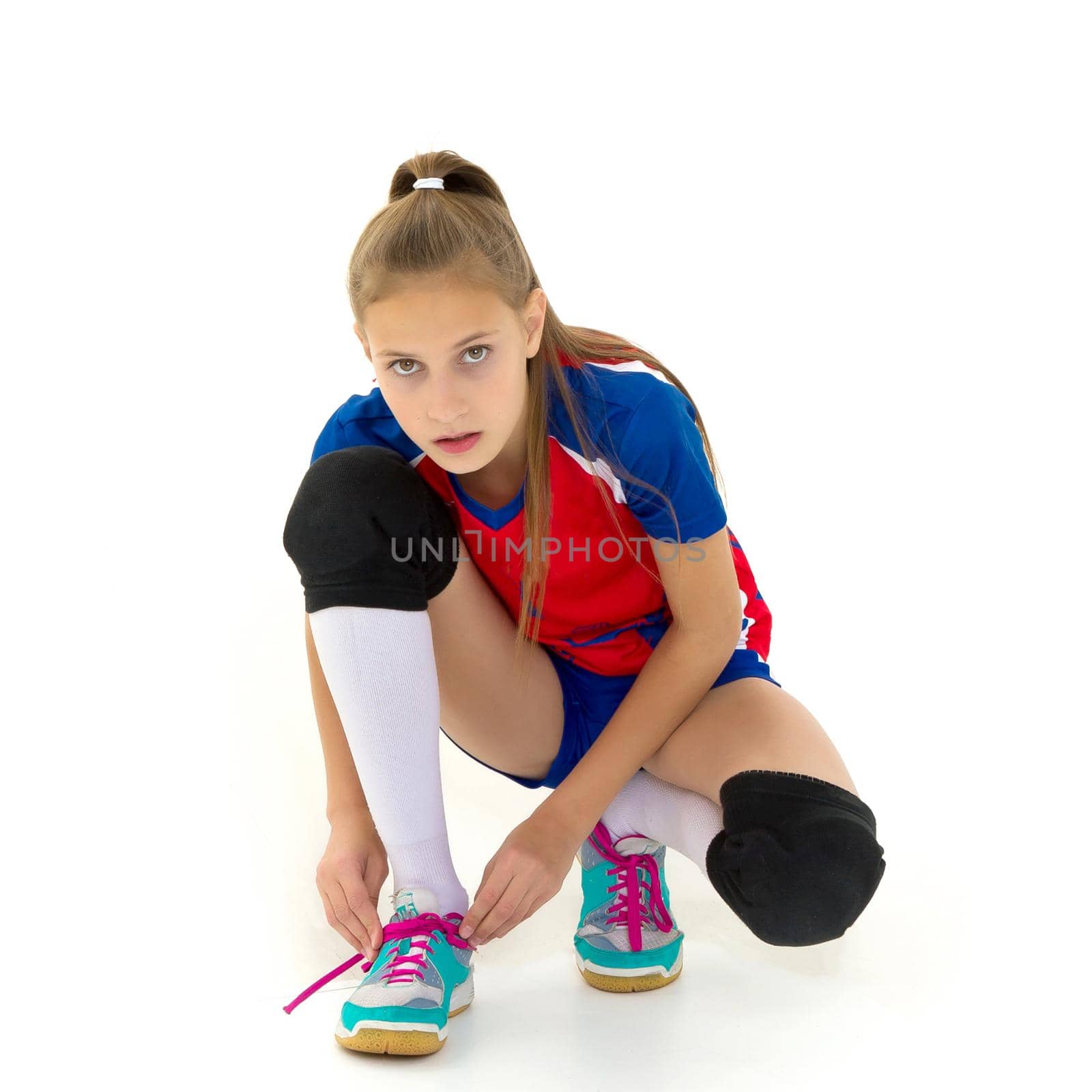 Girl crouching down and tying her shoelaces. Pretty teenage girl volleyball player in sports uniform, knee pads and sneakers posing on isolated white background looking at camera
