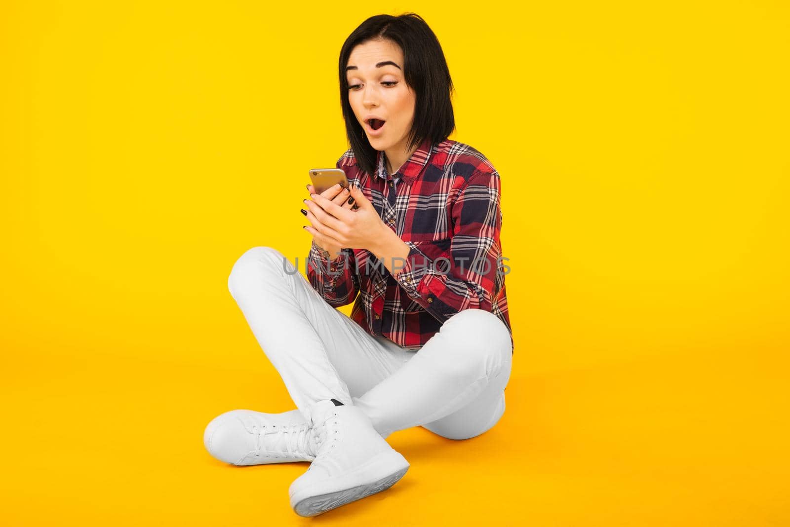 Excited laughing woman in plaid shirt sits and using mobile phone over yellow background - Image