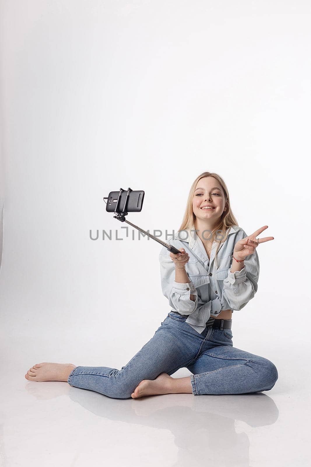 happy female making self portrait with smartphone attached to selfie stick. young pretty blond girl with long hair smiling, using cell phone isolated on white background, showing peace sign gesture