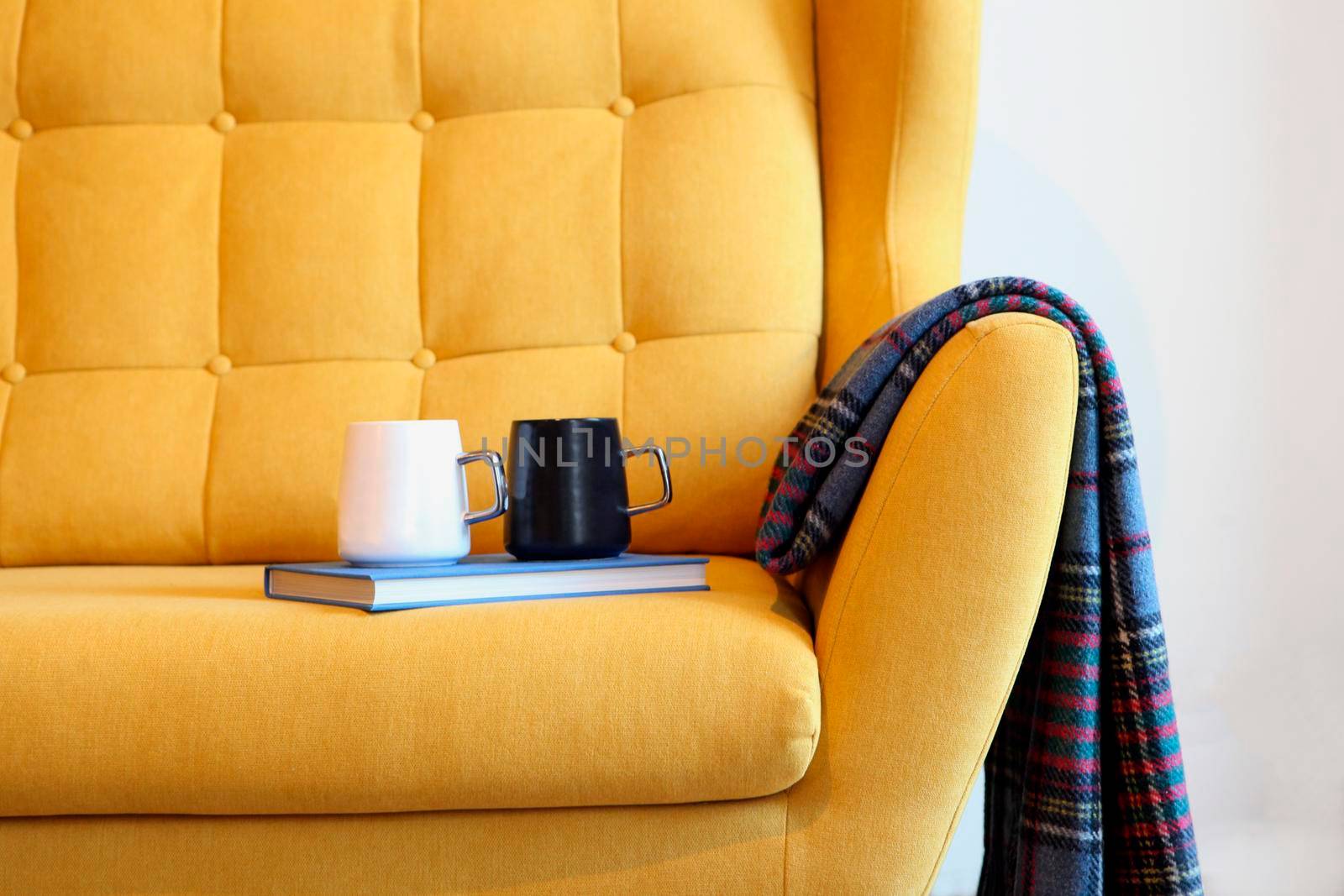 Cup of tea and blue book on a yellow coach with blanket. Still life details in home interior of living room. Cozy home interior, home comfort concept, gender free interior. Modern interior in the room.