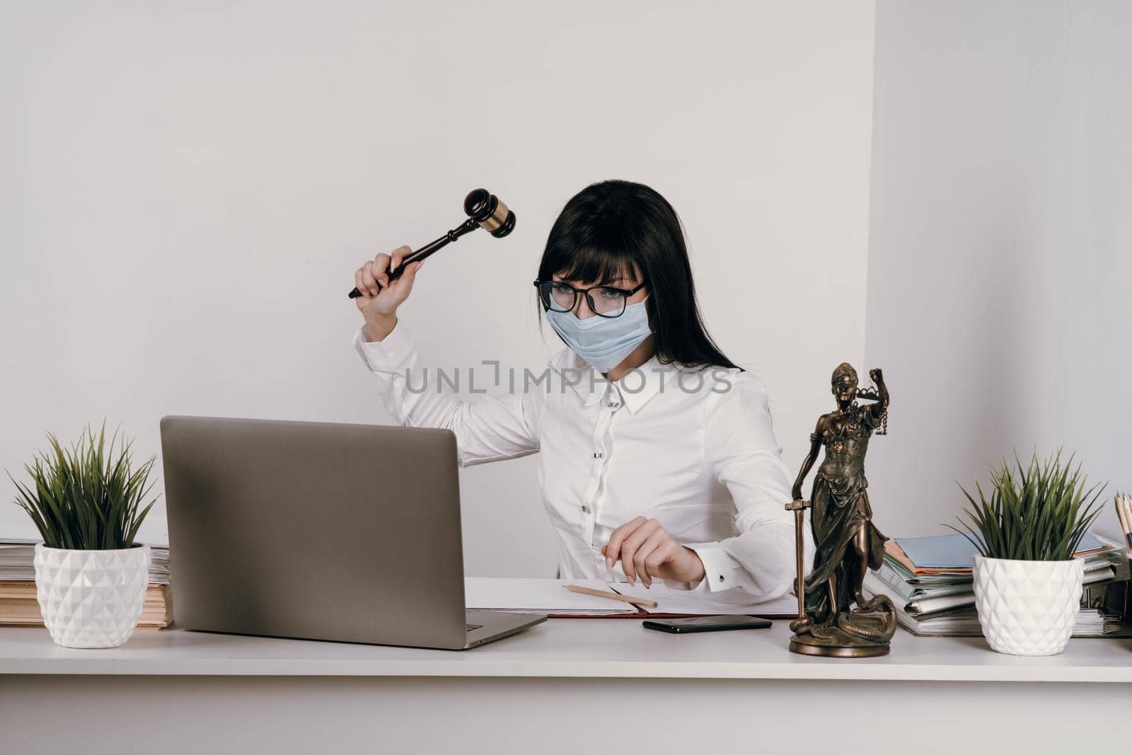 Young woman lawyer or judge works remotely in the office with a protective mask during an epidemic. Swings a hammer to pronounce judgment.