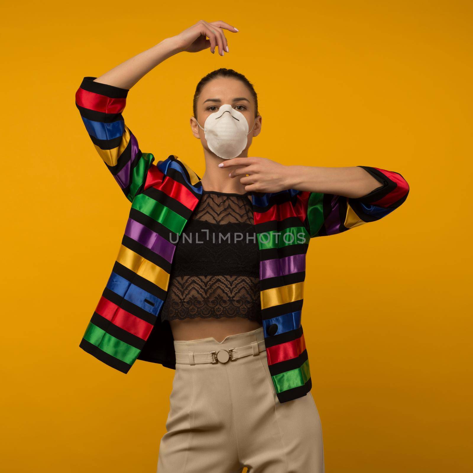 Beautiful slim girl fashion model posing in a protective respirator on a yellow background. LGBT community rainbow color jacket