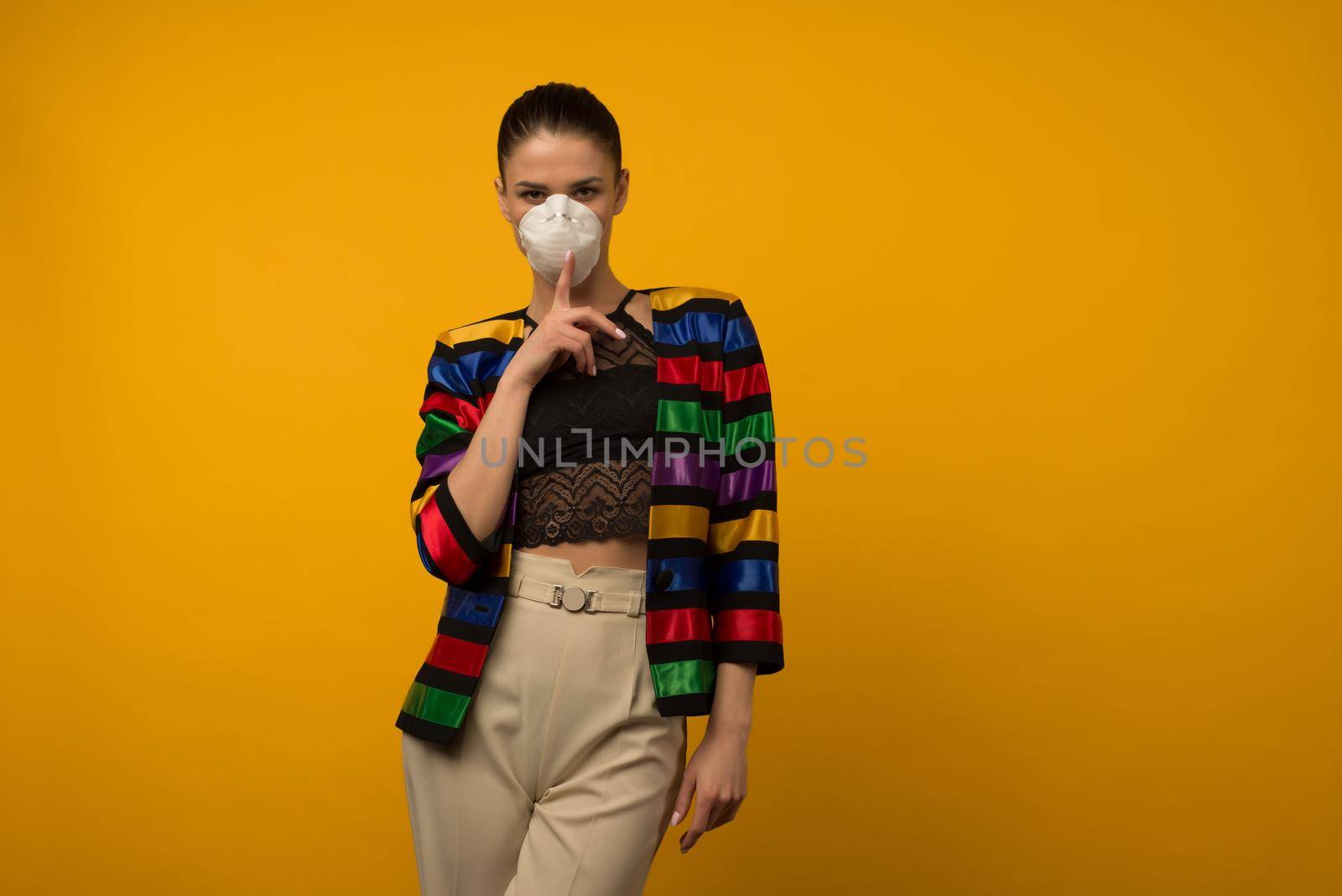 Beautiful slim girl fashion model posing in a protective respirator on a yellow background. LGBT community rainbow color jacket