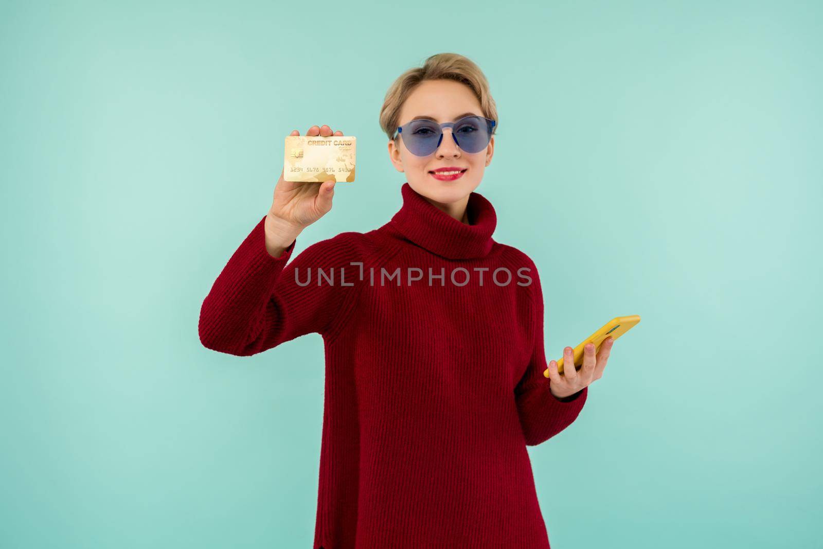 Portrait of beauty young girl in sunglasses showing plastic credit card while holding mobile phone isolated over blue background