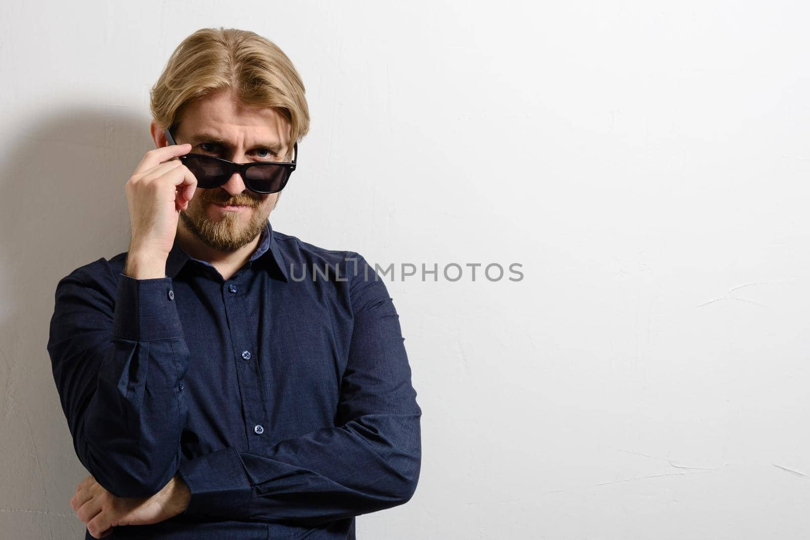 Stylish man in a blue shirt and sunglasses standing near a white wall and a hand holding glasses