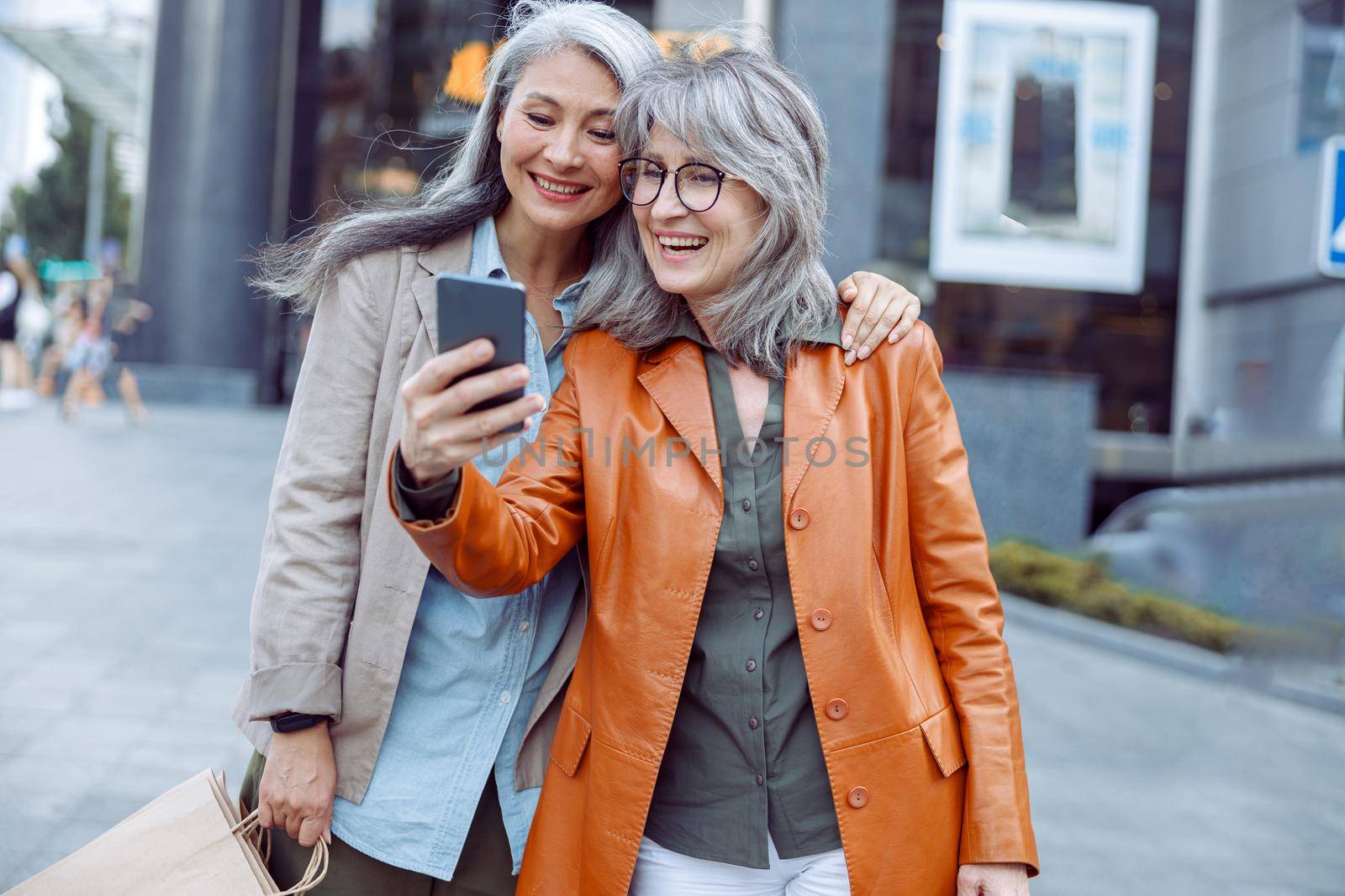 Smiling silver haired lady in brown leather jacket and companion with shopping bags take selfie on modern city street on autumn day. Friends spend time together