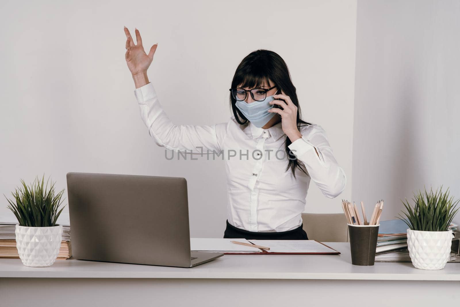 A young woman works remotely in the office with a protective mask during an epidemic. Conducts a video conference and talks on the phone