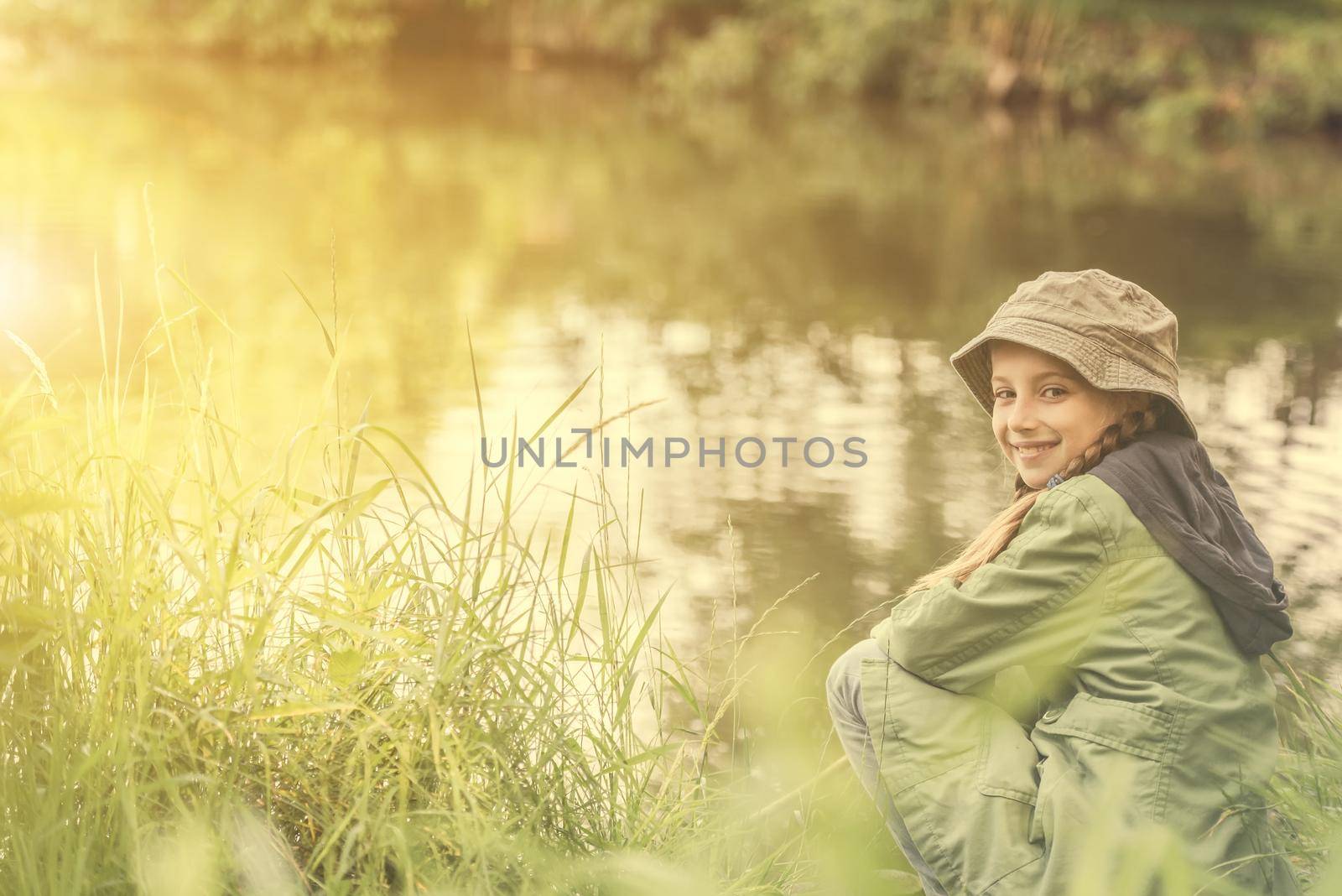 little fisher girl sitting on a river bank with a rod smiling