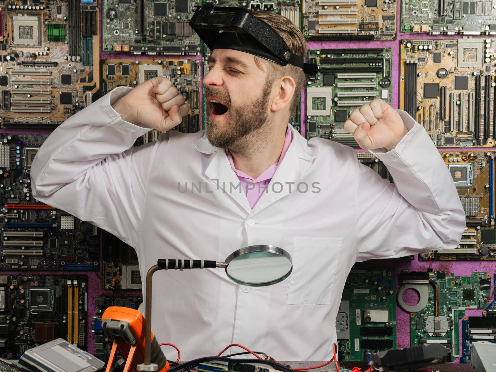 Young male electrician engineer tiredly yawns while sitting at the table of the electrical laboratory.