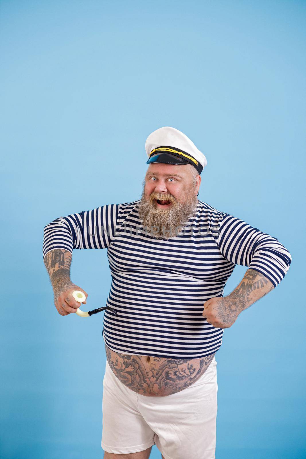 Funny dashing bearded plus size person wearing sailor costume with smoking pipe poses on light blue background in studio