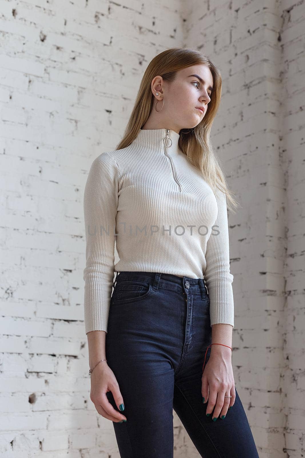 portrait of cute thoughtful teen model wearing white sweater and black jeans. caucasian skinny young female with blond hair stands in front of white brick wall. natural pretty lady standing at studio