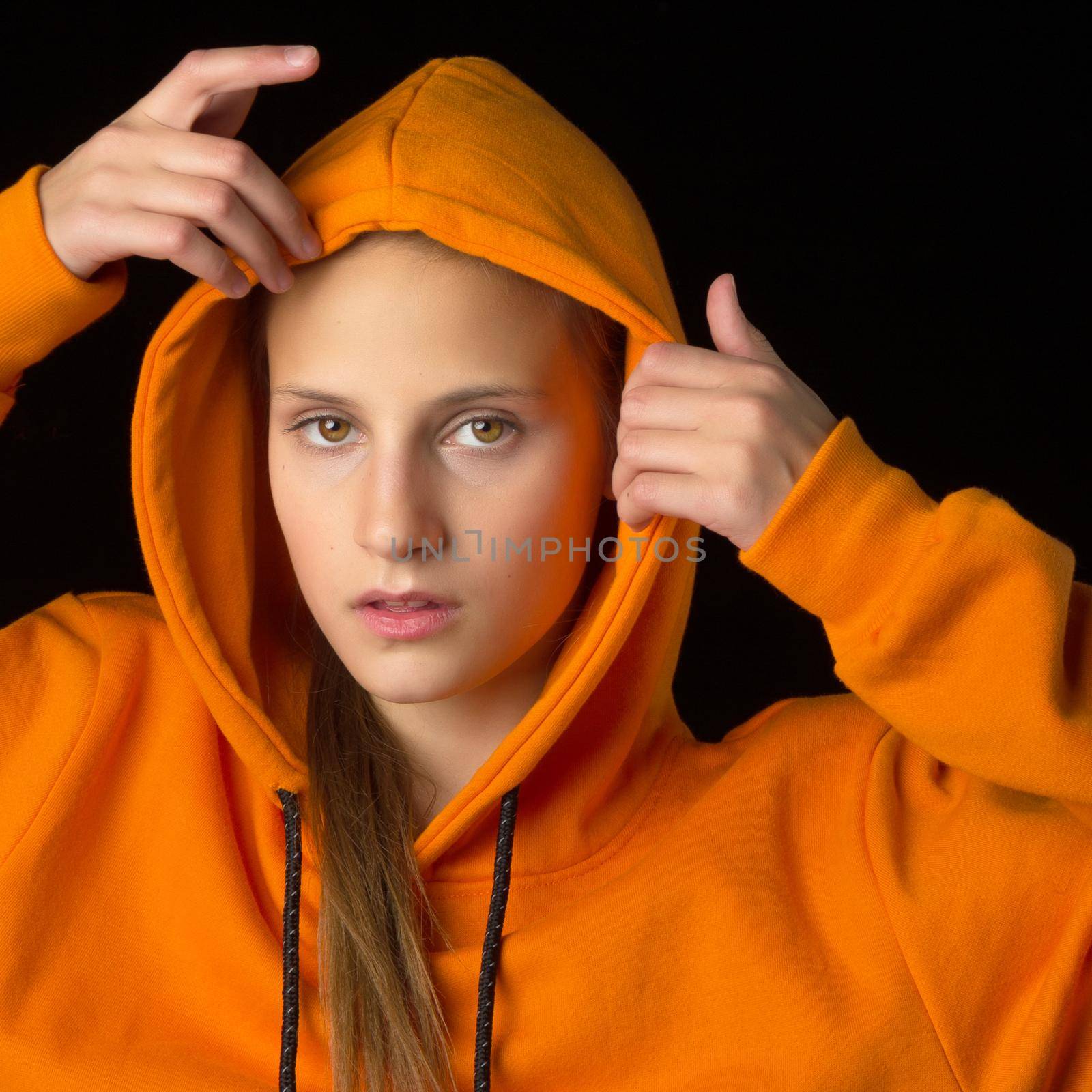 Shot of pretty teenage girl putting the hood on. Portrait of attractive teenager wearing warm orange hoodie standing against black background. Beautiful brown eyed girl smiling at camera