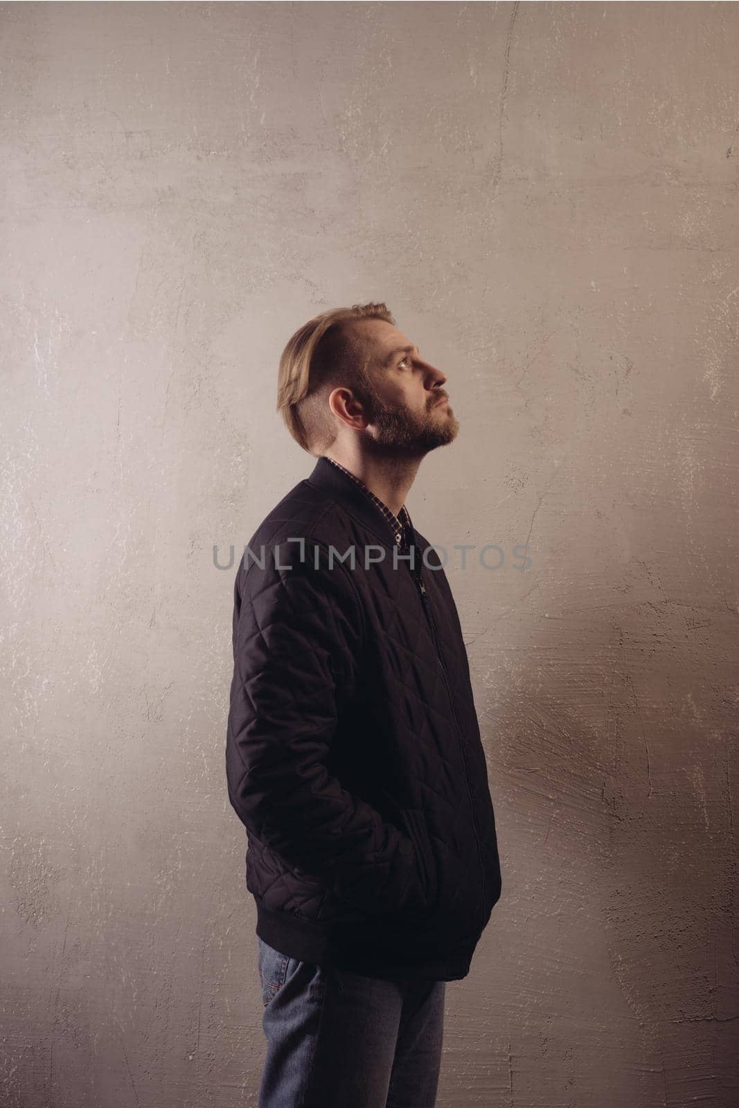 An attractive middle-aged bearded man in a black jacket and jeans stands behind the wall looking sideways to the top. copy space. deadpan without retouching