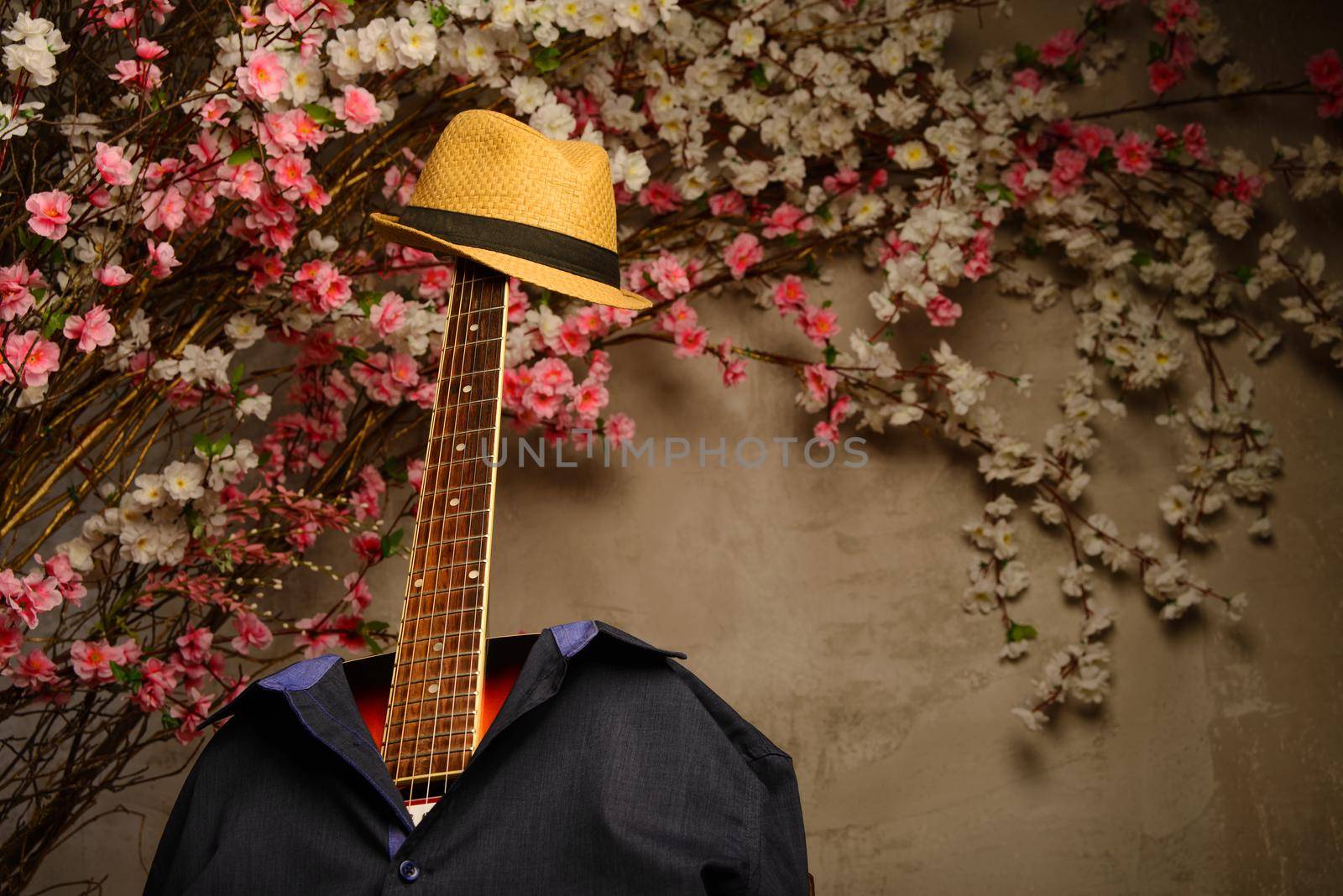 guitar is dressed in a man's blue shirt and a hat with a fretboard near a wall of flowers.