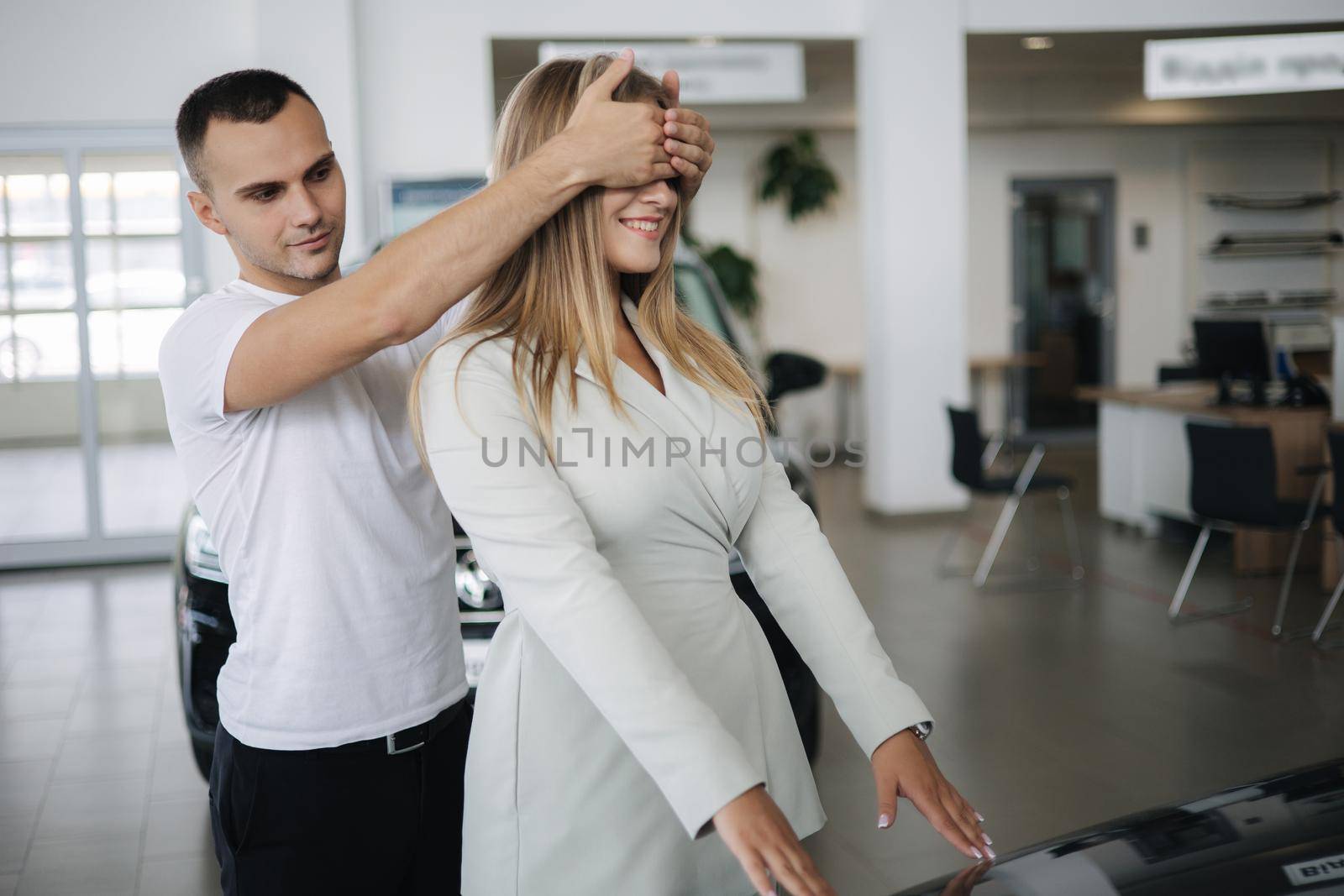 Man close eyes to his wife and makes a surprise buying car. Man and woman in car showroom by Gritsiv