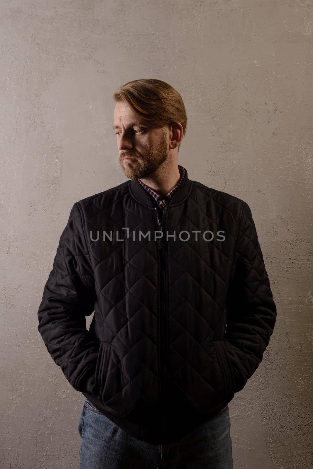 Cute middle-aged bearded man in a black jacket and jeans stands behind the wall looking away. copy space. deadpan without retouching