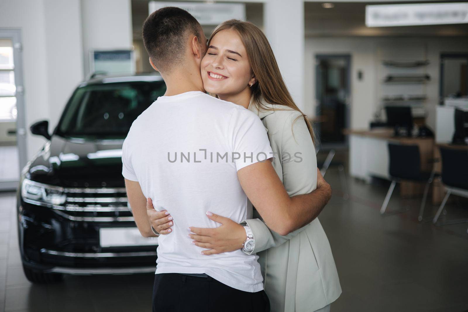 Happy woman hug his husband afrer buying car in car showroom. Man and woman buy new car.