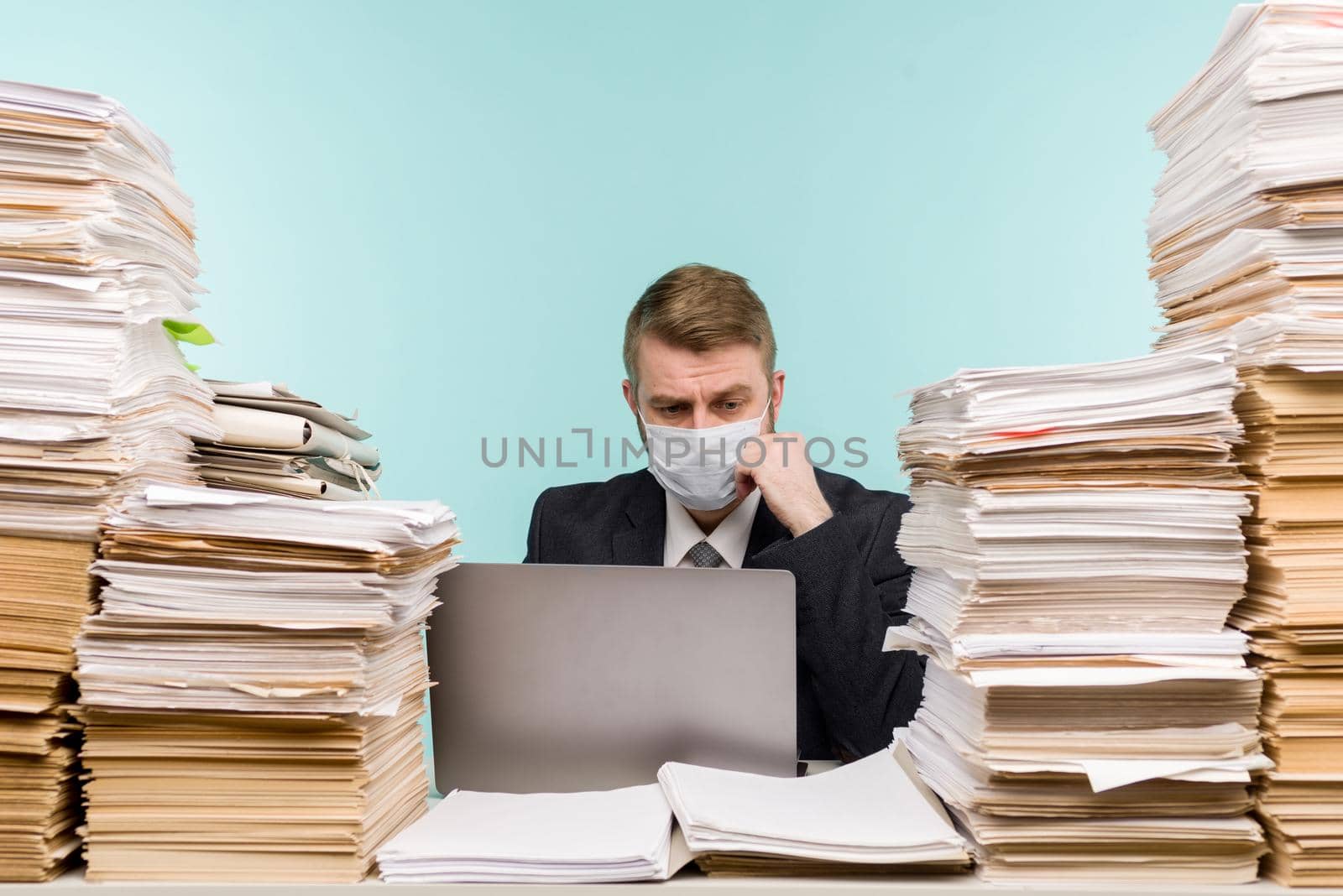 A male accountant or company manager works in an office in a pandemic in view of the accumulated paper work. A protective medical mask is on the face. On the desktop are large stacks of documents. - image
