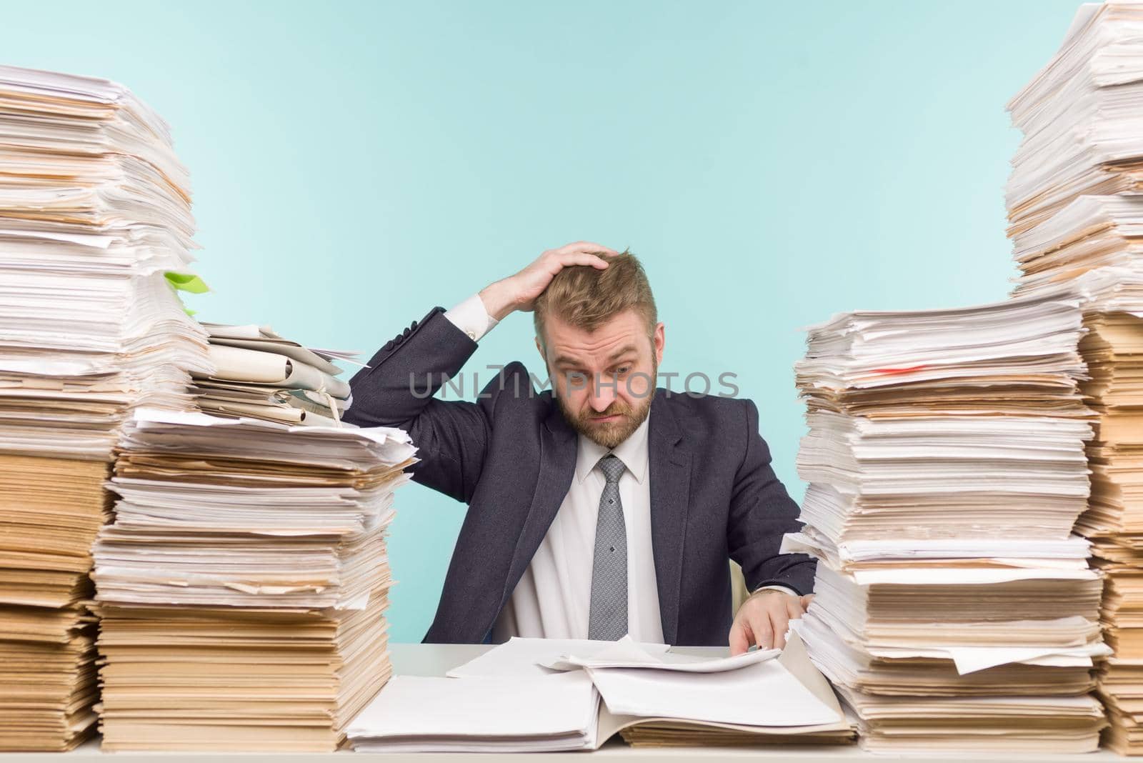Shocked businessman sitting at the table with many papers in office, he is overloaded with work - image