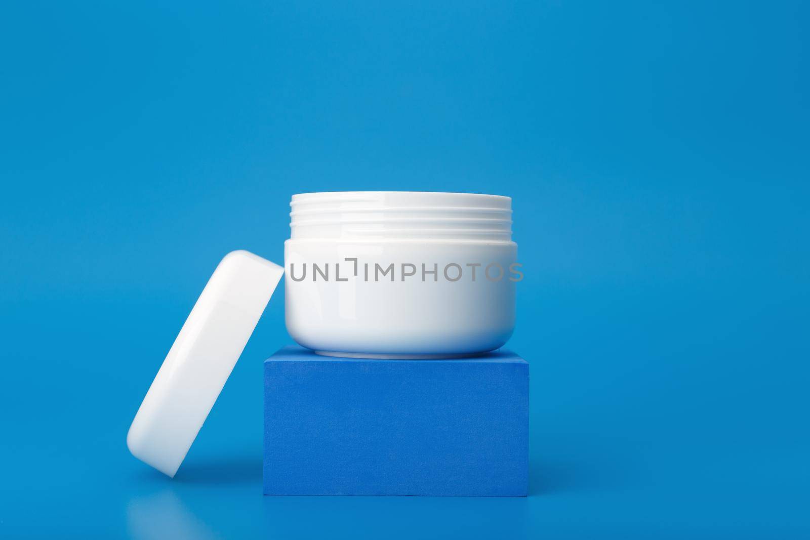 Minimal still life with white opened cosmetic jar on pedestal against dark blue background. Concept of repairing, moisturizing, anti aging nourishing night cream, gel, mask or lotion
