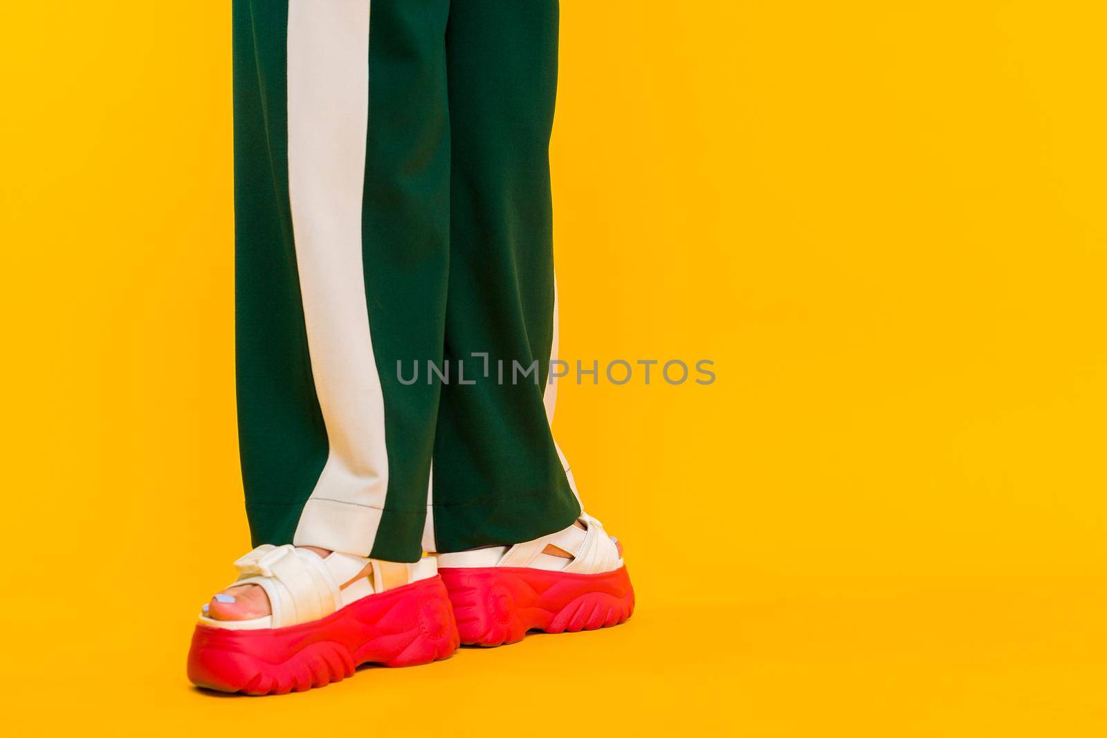 Women's legs in sport sandals with red soles and green pants with stripes on a yellow background.