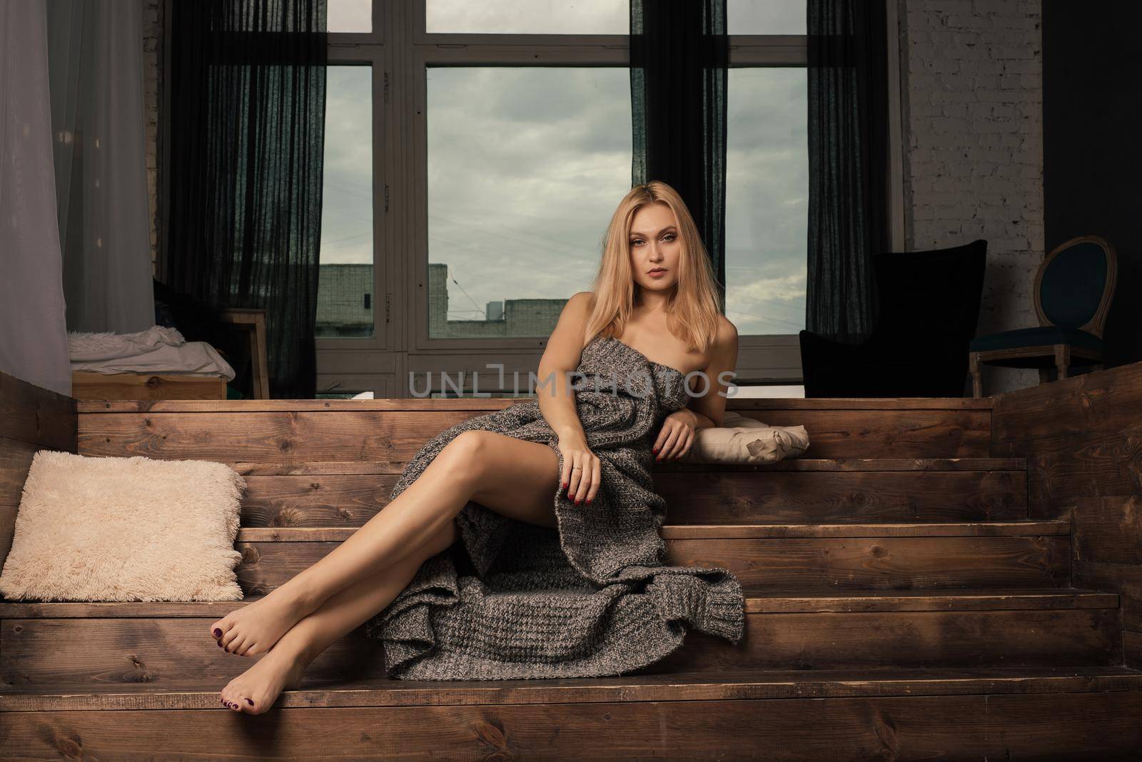 Beautiful young blond woman lies on a wooden staircase wrapped up in a woolen blanket.