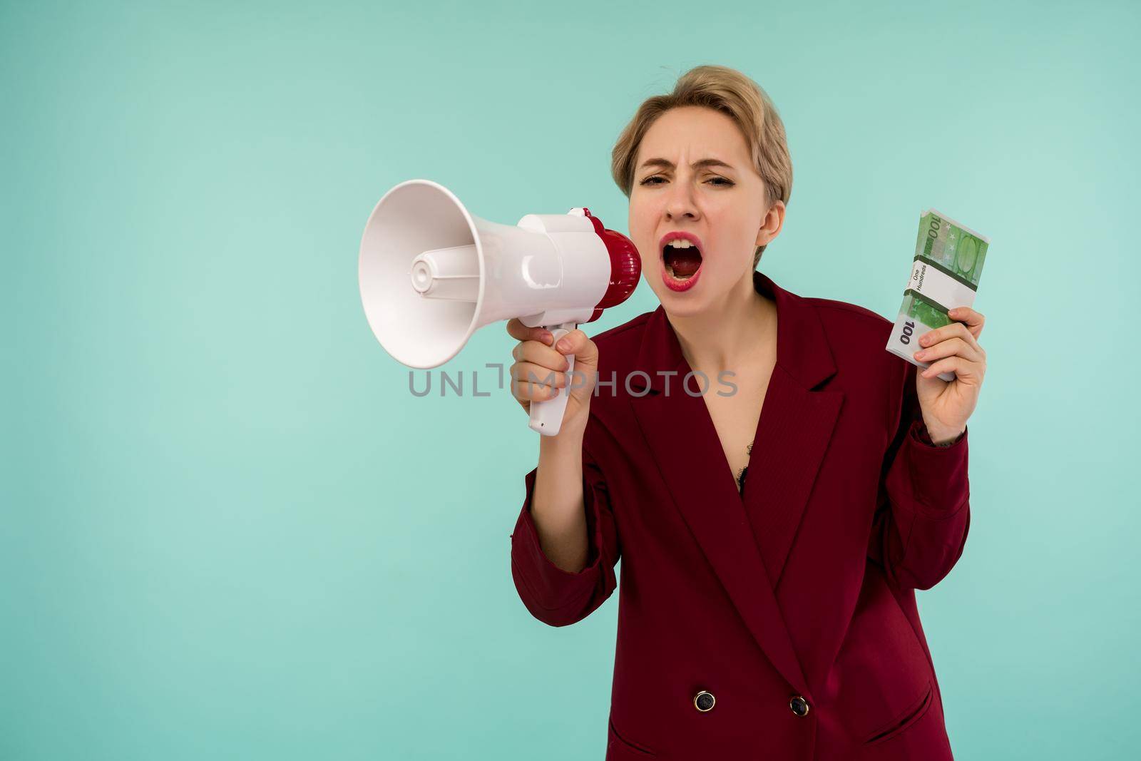 Portrait of beautiful young screaming businesswoman with money and megaphone, on blue background - Image