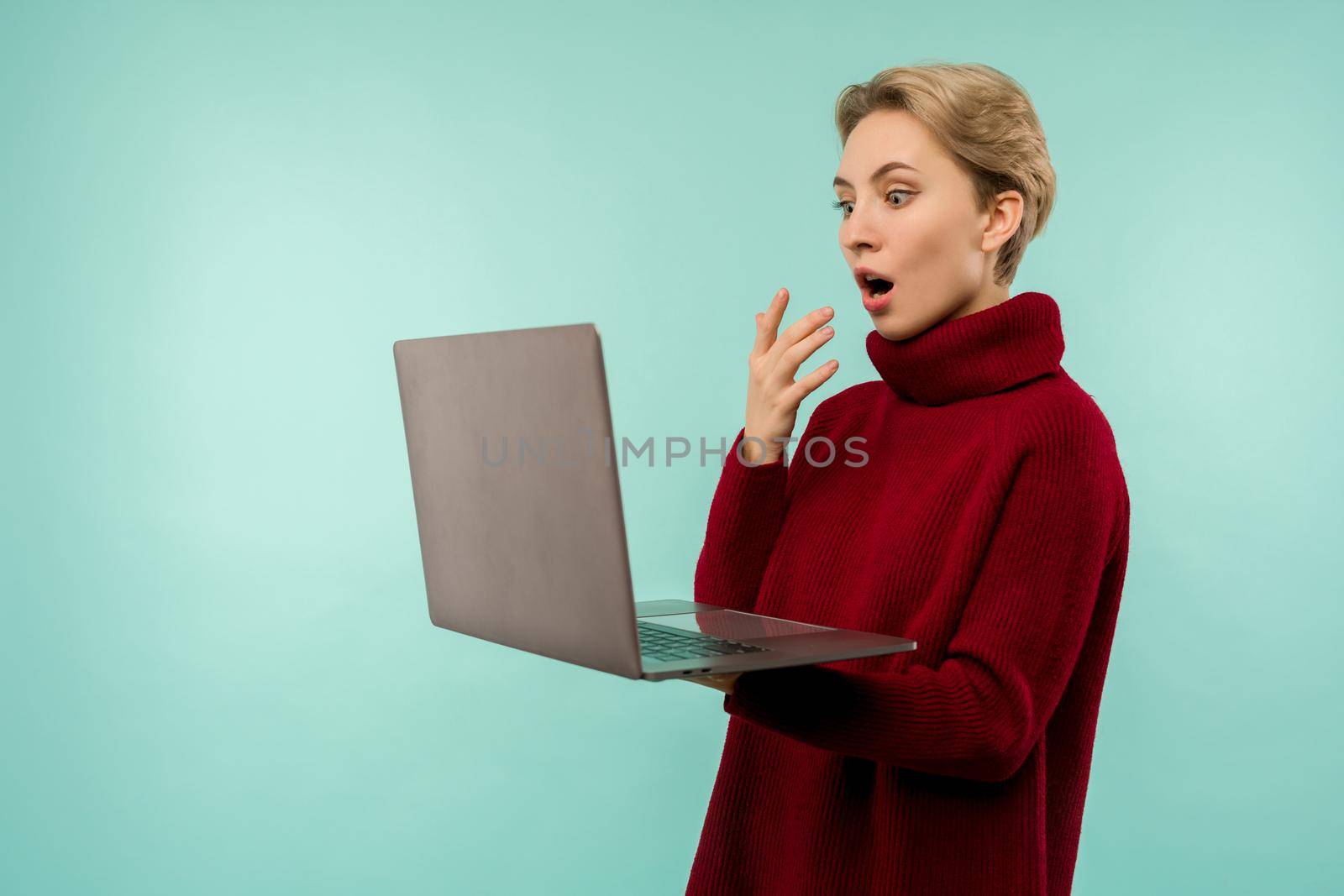 Surprised happy blonde woman in sweater looking laptop computer screen with open mouth over blue background by zartarn