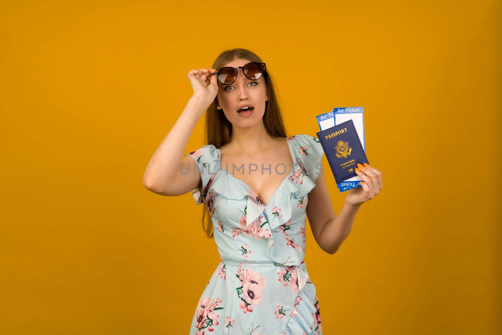 Pop-eyed young woman in blue dress with flowers and sunglasses is holding airline tickets with a passport on a yellow background. Rejoices in the resumption of tourism after the coronovirus pandemic.