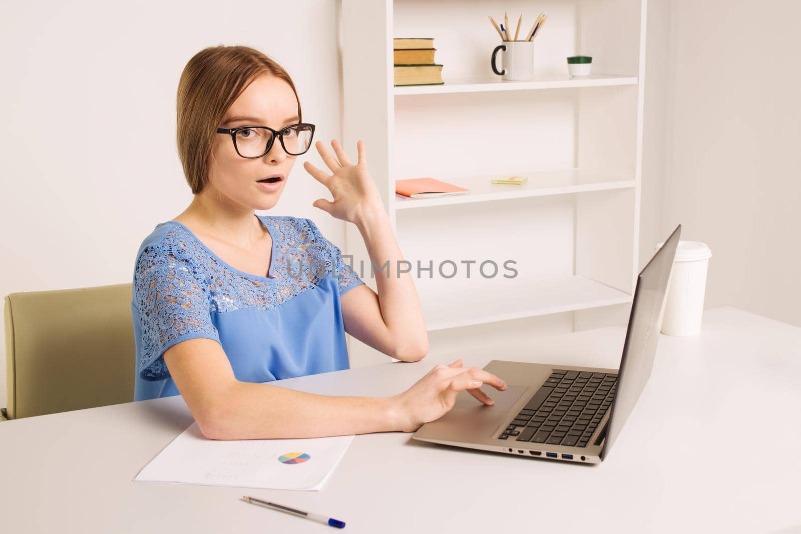 Portrait of an attractive woman at the table with cup and laptop, book, notebook on it, grabbing her head. Bookshelf at the background, concept photo - Image