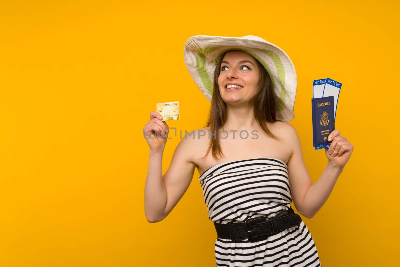 Joyful young woman in a straw hat and a striped dress is holding airline tickets with a passport and a credit card on a yellow background. Rejoices in the resumption of tourism after the coronovirus pandemic.