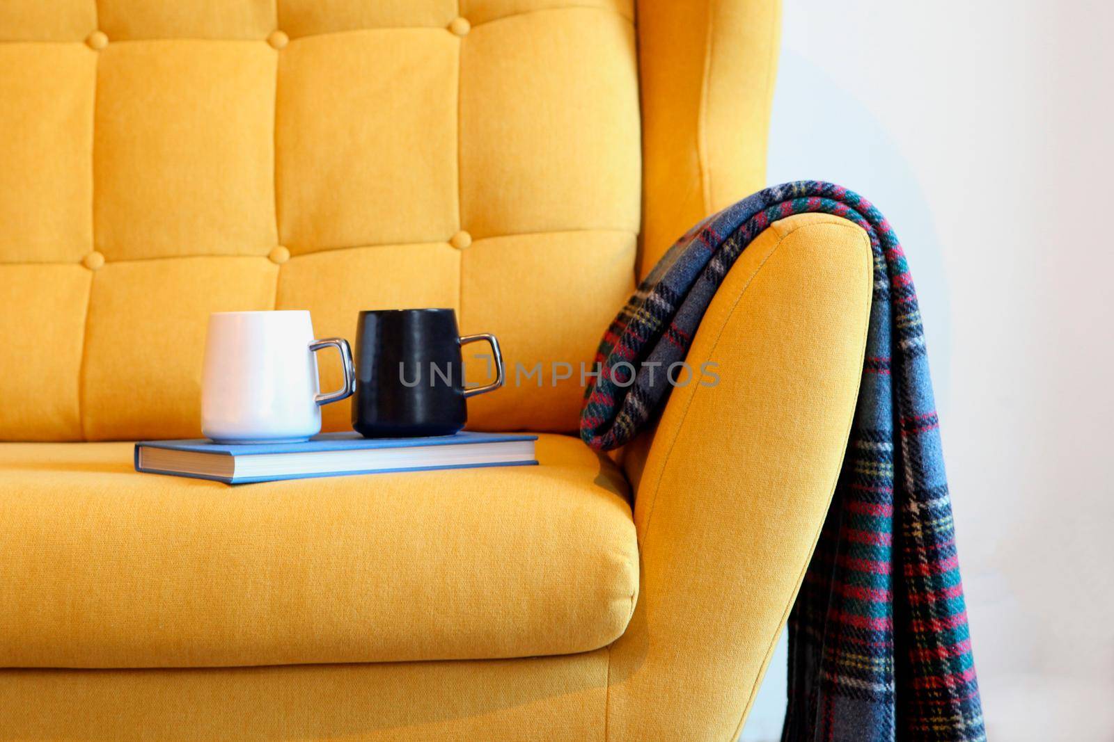 Cup of tea and blue book on a yellow coach with blanket. Still life details in home interior of living room. Cozy home interior, home comfort concept, gender free interior. Modern interior in the room.