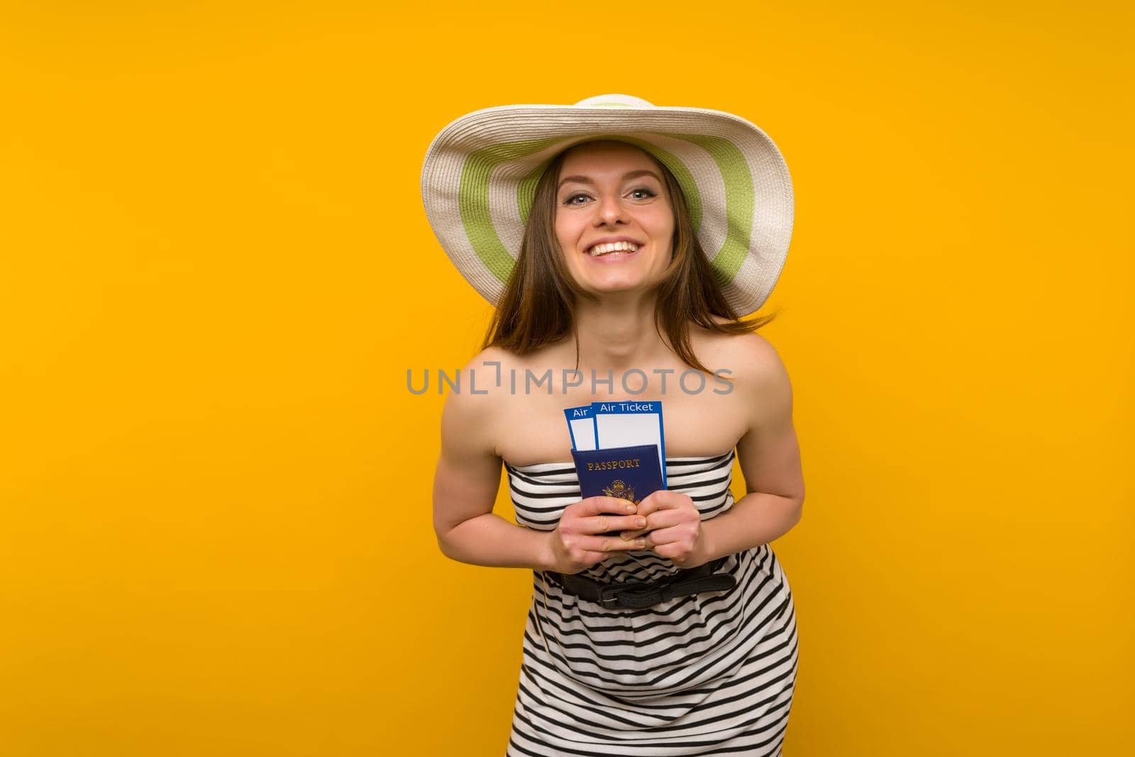 Joyful young woman in a straw hat and a striped dress is holding airline tickets with a passport on a yellow background. Rejoices in the resumption of tourism after the coronovirus pandemic.