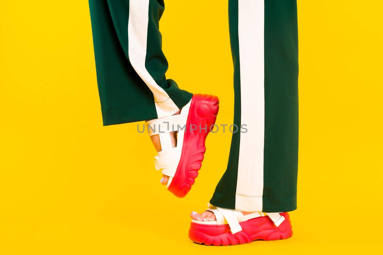 Women's legs in sport sandals with red soles and green pants with stripes on a yellow background.