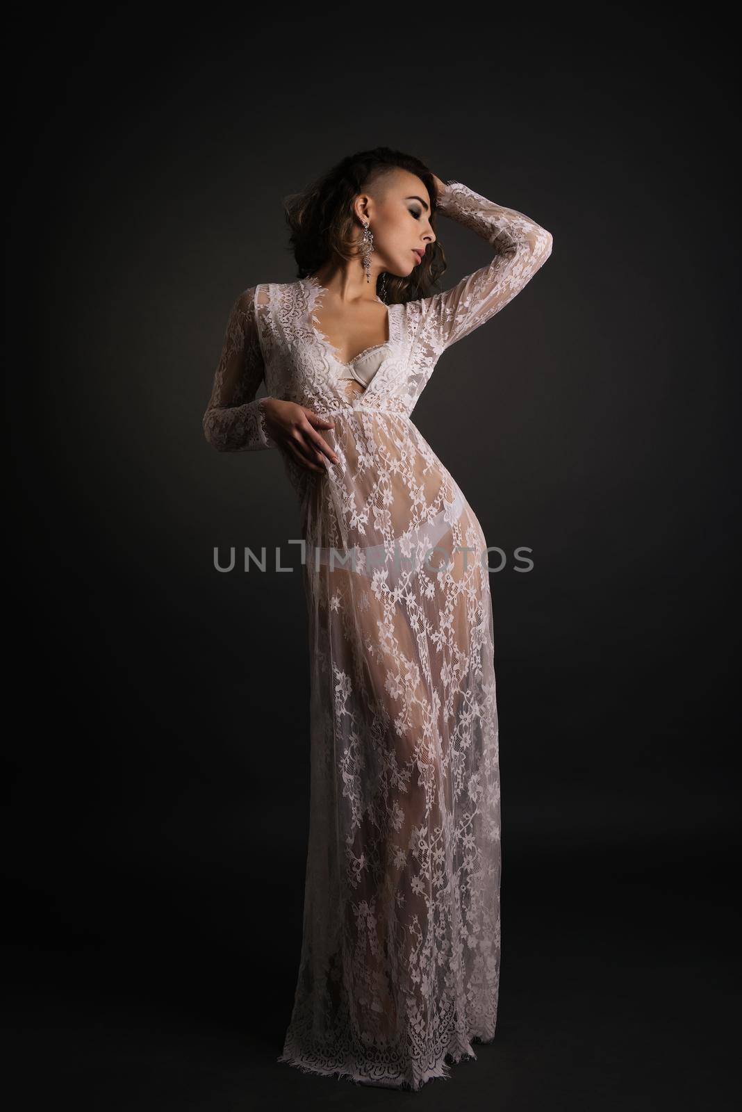 Young slim woman posing in white lace transparent dress on dark background