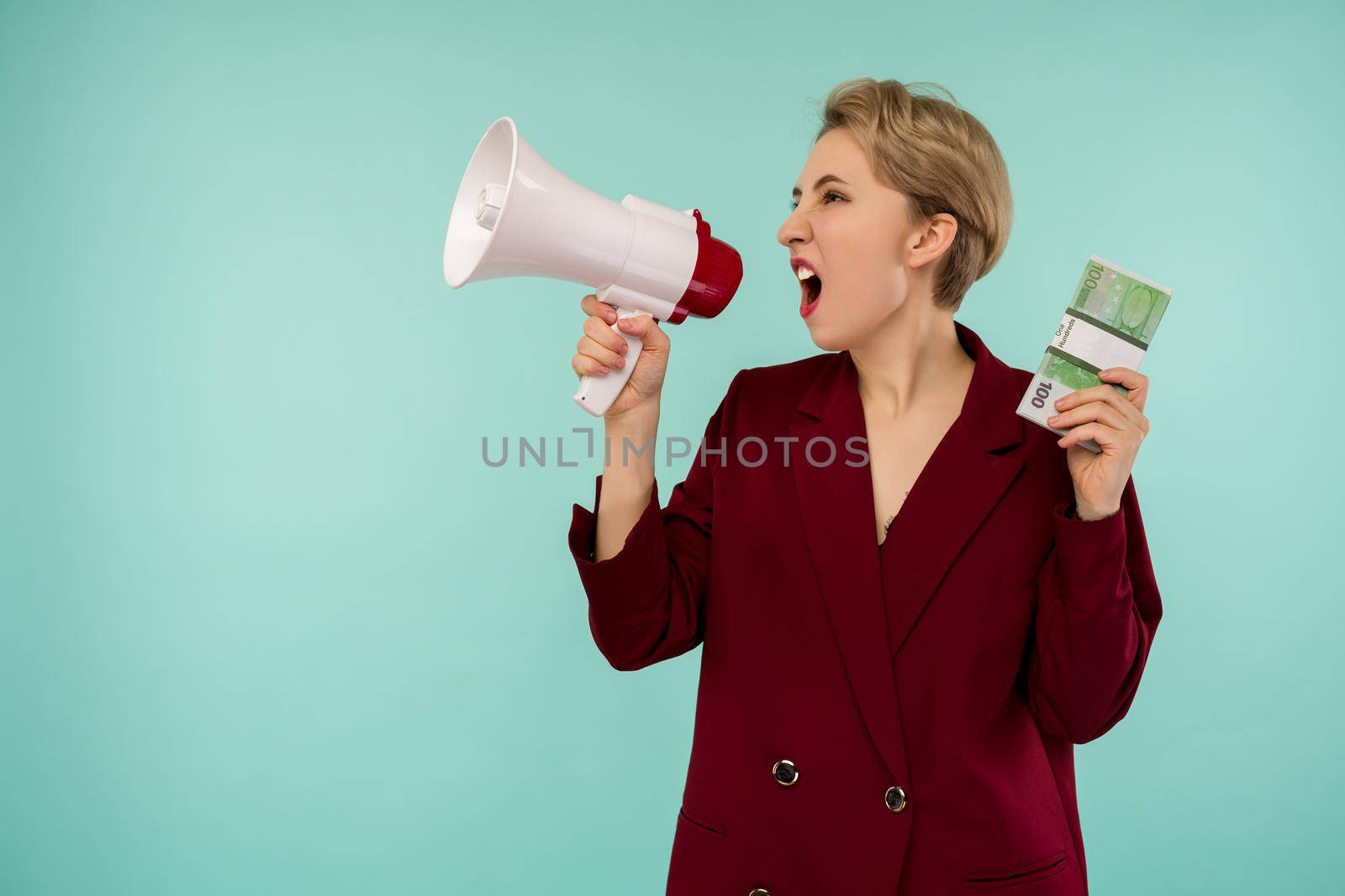 Portrait of beautiful young screaming businesswoman with money and megaphone, on blue background - Image
