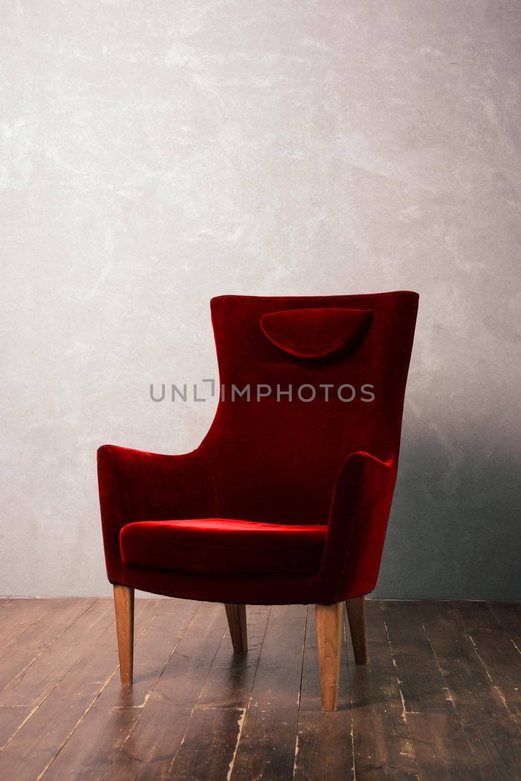 Luxurious red velor armchair stands near the textural wall on a dark wooden floor.