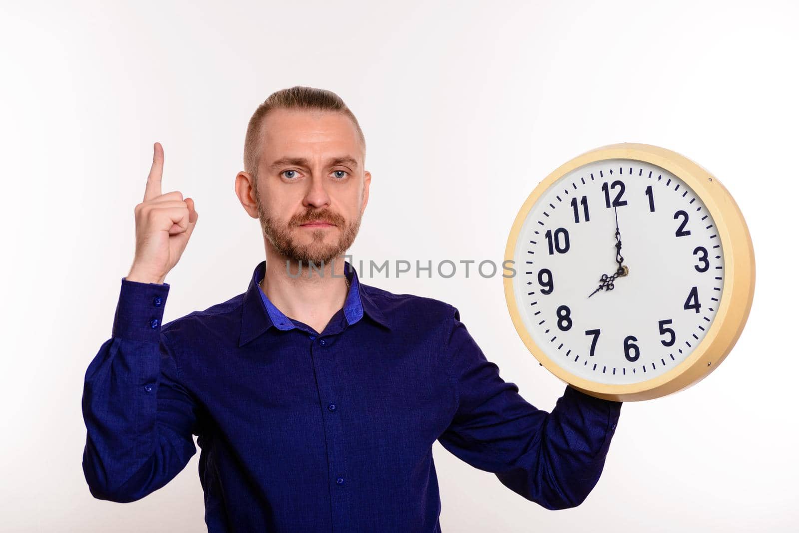 Young handsome man in blue shirt with big clock standing pointing upwards looking into the camera on white background.