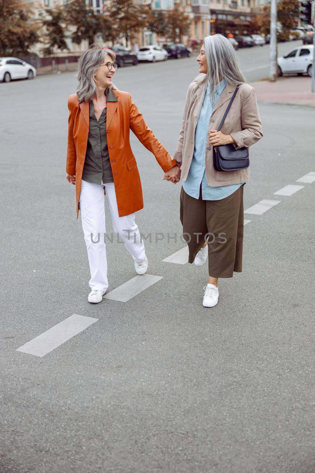 Happy senior woman with Asian companion walk joining hands along contemporary city street on autumn day. Friends spend time together